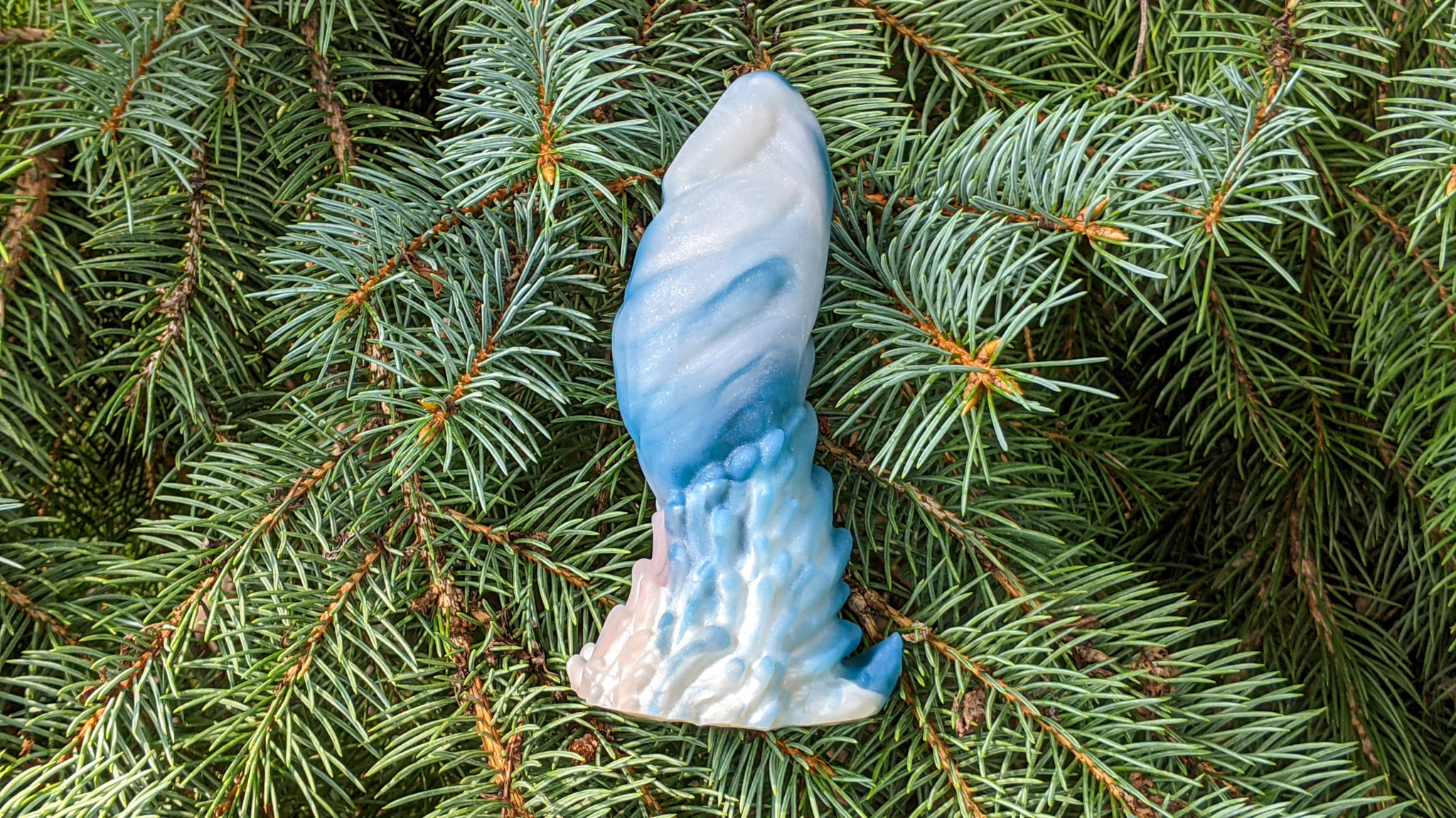 Ceela is a blue and white fantasy dildo laying on its side. In this image, it's resting on top of some fir tree branches.